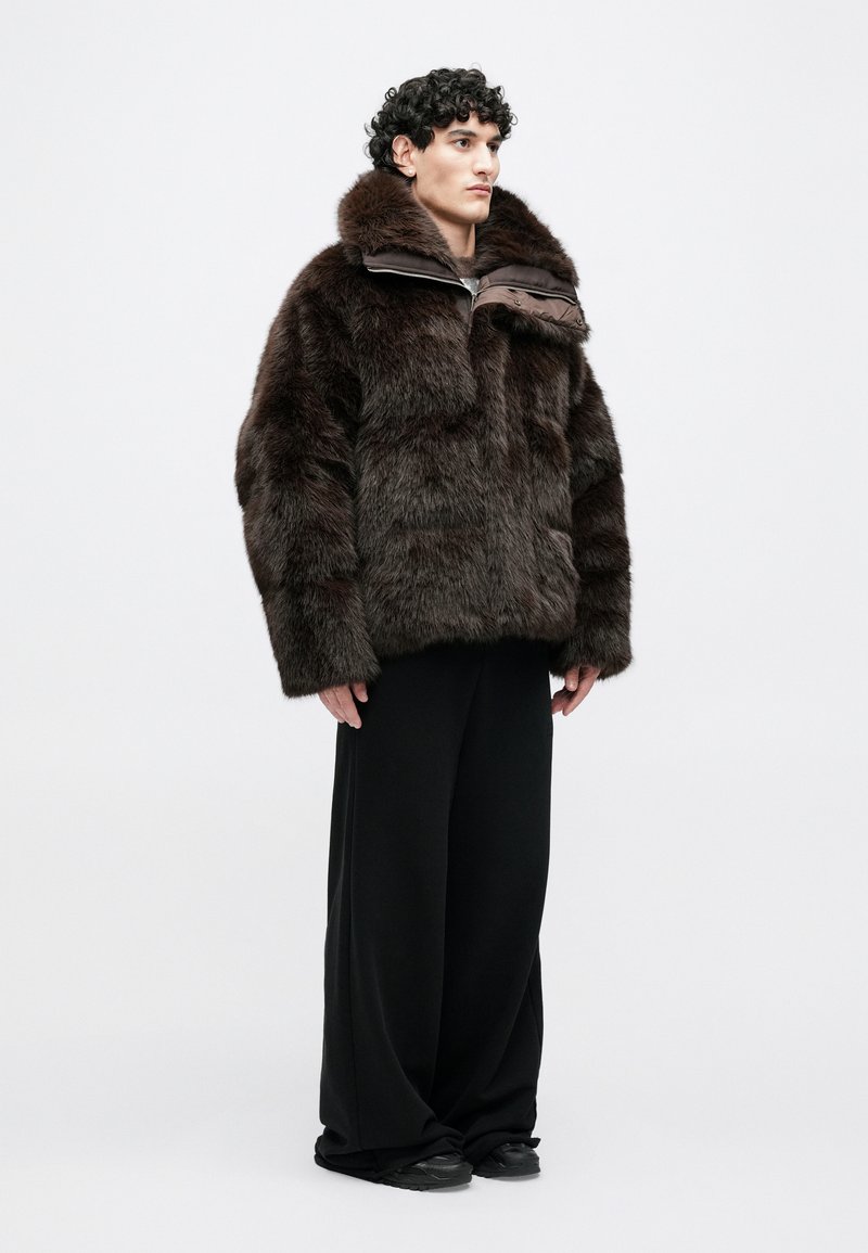 Brown fur jacket with a high collar and dual-tone front, paired with loose black pants and black footwear. Soft texture, voluminous design.