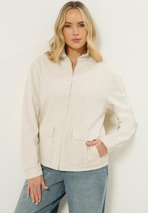 Lyshåret kvinde iført beige lynlåstrøje med klaplommer og blå jeans, stående foran en ensfarvet lys baggrund.