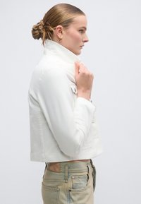 Λευκό cropped μπουφάν με ψηλό γιακά και μπροστινές τσέπες, κατασκευασμένο από απαλό ύφασμα, συνδυασμένο με ανοιχτόχρωμο μπλε τζιν, παρουσιάζοντας έναν μινιμαλιστικό σχεδιασμό.