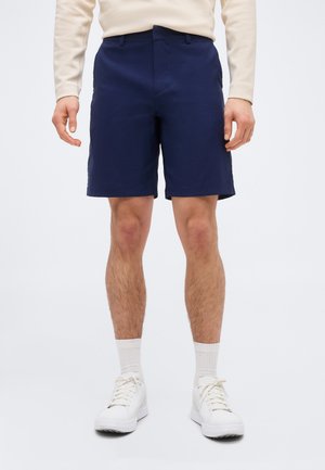 Hombre con camiseta de manga larga beige, pantalones cortos azul marino, calcetas blancas y zapatillas blancas, de pie contra un fondo blanco liso.