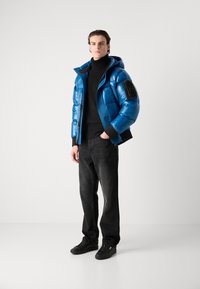 Zils pūkains mētelis ar spīguļojošu tekstūru, kapuci un melnu pielikumu, kopā ar melnu turtleneck un tumšiem džinsiem, pabeigts ar melniem sporta apaviem.