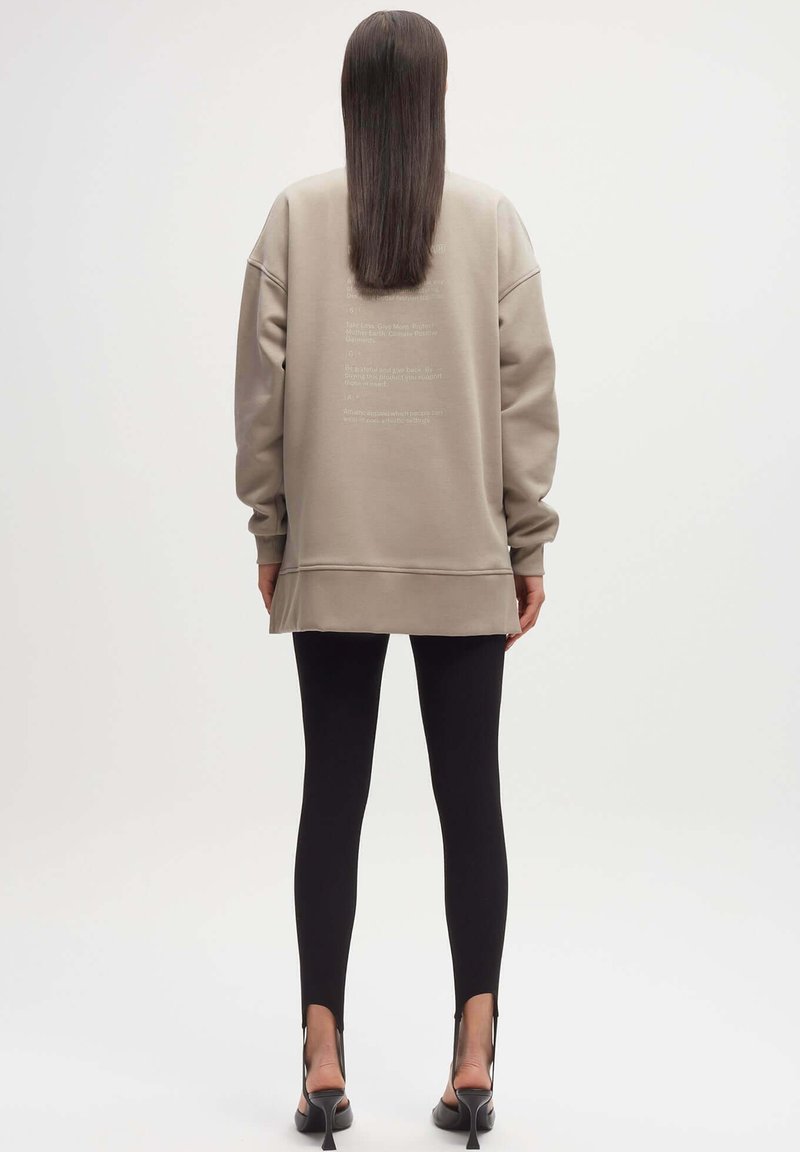 Overgroot beige sweatshirt met verlaagde schouders, ribgebreide manchetten en tekstdetails op de achterkant, gecombineerd met zwarte legging met sleuven bij de enkels.