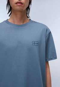 Camiseta de algodón azul claro con cuello redondo, mangas cortas y un pequeño logo de bandera impreso en la parte izquierda del pecho.