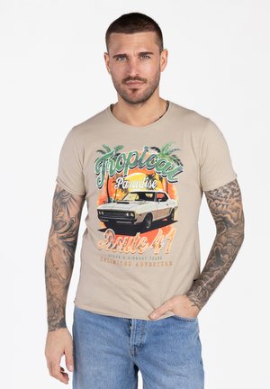 Mann mit tätowierten Armen trägt ein beiges T-Shirt mit einem Vintage-Auto, Palmen und dem Schriftzug "Tropical Paradise Route 41" sowie eine blaue Jeans.