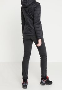 Doudoune noire avec capuche, design extensible et matelassé, associée à un jean slim noir et des chaussures noires avec des accents rouges.