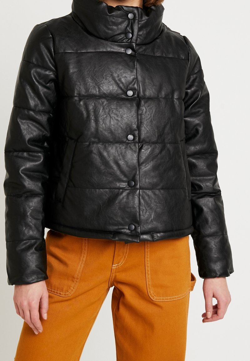 Veste noire matelassée avec boutons-pression portée sur un pantalon couleur moutarde avec grandes poches avant.