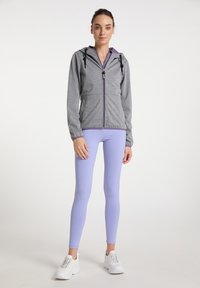 Szürke cipzáros kapucnis pulóver lila részletekkel, lila leggings-szel párosítva. A modell fehér sportcipőt visel, egy sima háttér előtt áll.