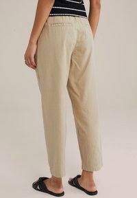 Light beige tapered pants with a slightly wrinkled texture, featuring a gathered waistband and back pockets, paired with black slide sandals.