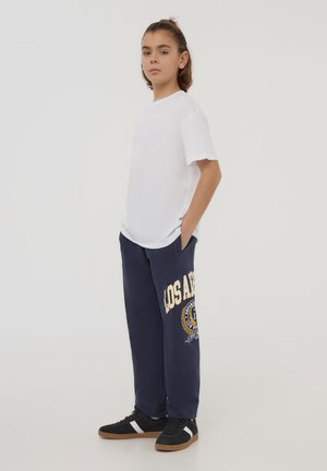 T-shirt in cotone bianco abbinato a pantaloni della tuta blu navy con grafica "LOS ANGELES" e dettagli a fantasia. Sneakers nere con righe bianche.