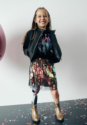 Ein Kind trägt eine schwarze Lederjacke über einem glitzernden, mehrfarbigen Rock, mit goldenen Knöchelstiefeln und einem sichtbaren Prothesenbein.