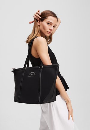 Black tote bag made of smooth material, featuring dual handles, a zip closure, and logo detailing on the front. Simple, structured design.