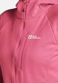 Veste légère rose avec une finition texturée, dotée d'une fermeture éclair à l'avant et d'une capuche, ainsi qu'un logo blanc sur la zone gauche de la poitrine.