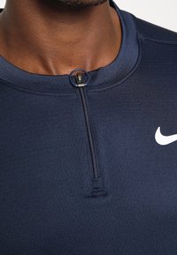 Tummansininen urheilupaita, jossa on teksturoitu kangas, lyhyt vetoketju kauluksessa ja valkoinen Nike-logo vasemmassa rintakappaleessa.
