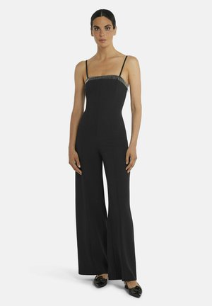 Vrouw staat, draagt een aansluitende zwarte jumpsuit met dunne bandjes en wijd uitlopende broekspijpen, gecombineerd met zwarte puntige schoenen.