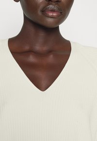 V-ringad topp i krämfärgad ribbad tyg, med subtil textur och tight passform. Fokus på neckline och mjuk hud.