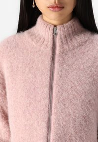 Suéter ligero de lana de color rosa con cremallera, cuello alto y acanalado, que cuenta con una textura suave y esponjosa, y un ajuste holgado.