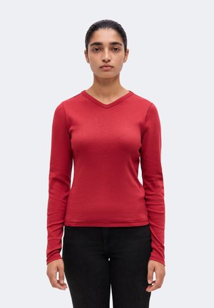 Femme aux cheveux foncés tirés en arrière, portant un t-shirt rouge à manches longues avec un col en V et un pantalon noir, debout devant un fond clair uni.