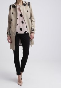 Trench coat beige con dettagli in pelle nera, bottoni doppi e vita con cintura. Indossato sopra un top rosa con motivi neri e pantaloni neri.