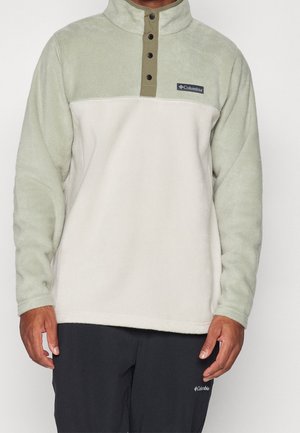 Homme portant un pull polaire Columbia vert clair et blanc cassé avec boutons-pression et pantalon noir Columbia, debout sur fond uni.