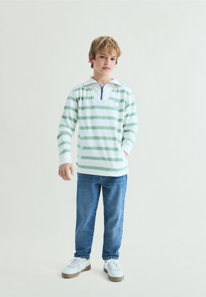 Chlapec stojící s rukama částečně v kapsách, v zeleno-bíle pruhovaném tričku s dlouhým rukávem, modrých džínách a bílých teniskách na neutrálním pozadí.
