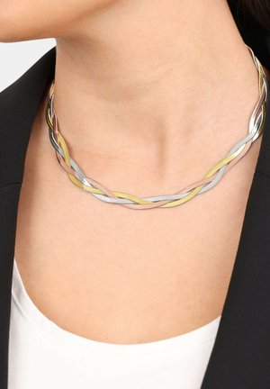 Primo piano di una persona che indossa una collana con catena a tre colori attorcigliata, con fili d'argento, oro e oro rosa, sopra una maglietta bianca e una giacca nera.