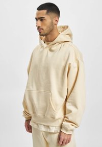 Beige oversized hoodie lavet af blødt stof, med en forlommer, en hætte med snoretræk og ribbede manchetter. Glat tekstur, minimalistisk design.