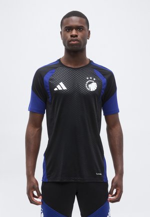 Homme portant un maillot de football noir et bleu du FC Copenhague avec le logo Adidas et l'emblème du lion, debout devant un fond blanc uni.