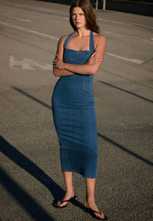Femme aux bras croisés portant une robe midi en denim bleu sans manches et des tongs noires, debout sur un sol pavé au soleil.