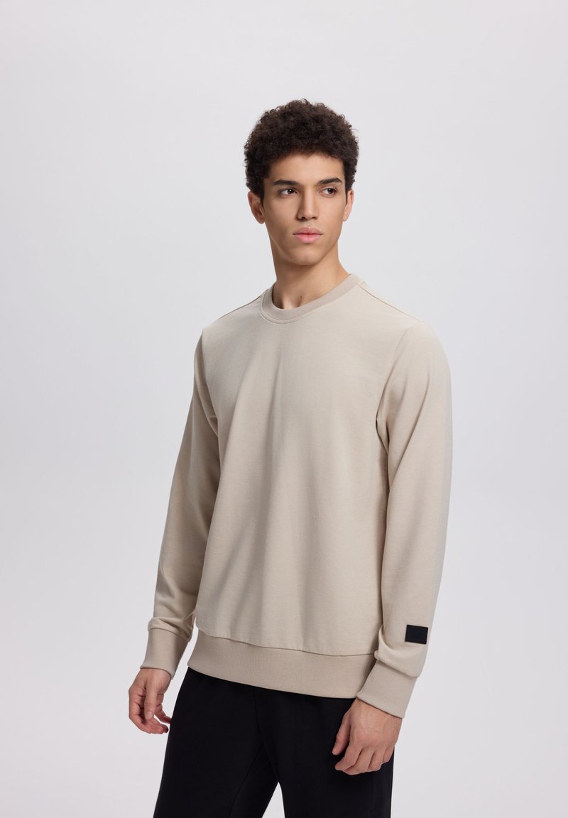 Giovane uomo che indossa una felpa beige a maniche lunghe e pantaloni neri, in piedi contro uno sfondo grigio chiaro uniforme, che guarda di lato.