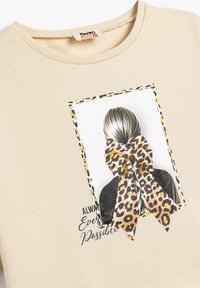 Sudadera beige con una impresión fotográfica de una persona con una trenza que lleva un lazo de estampado de leopardo. El texto dice "Siempre Todo es Posible."