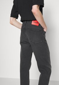 Jeans in denim nero con vestibilità dritta, con un'etichetta in pelle rossa sul retro. Cinque tasche, texture sottile e design pulito.