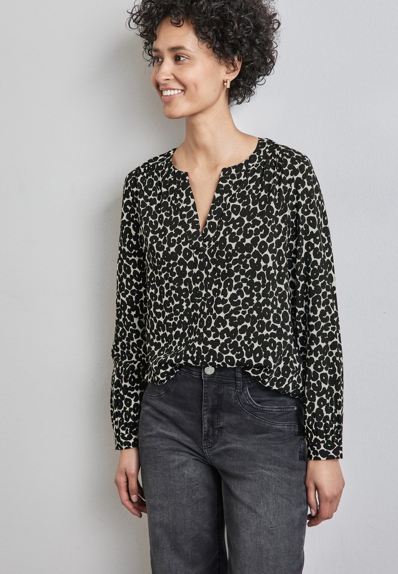 Blusa con stampa leopardata nera e beige, con scollatura a V e maniche lunghe, abbinata a jeans grigi scuri. Il tessuto appare liscio e leggero.