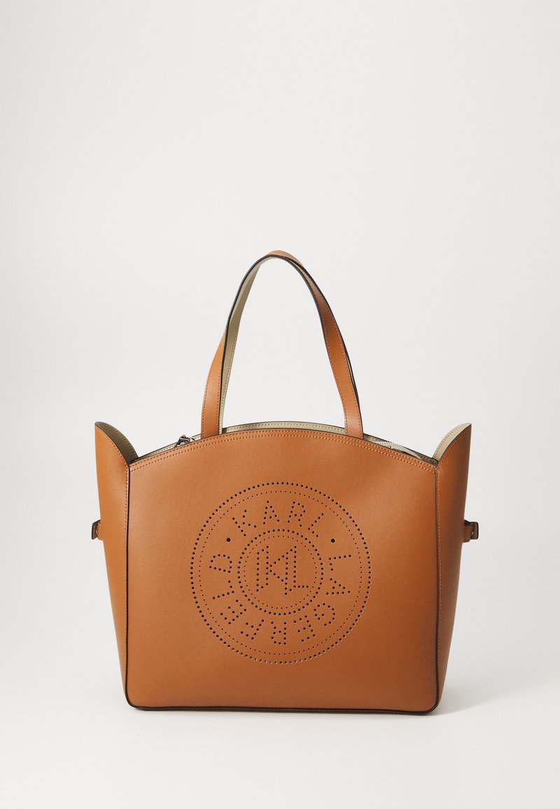 Sac cabas en cuir marron avec des anses arrondies et un logo circulaire perforé sur le devant, sur fond blanc uni.