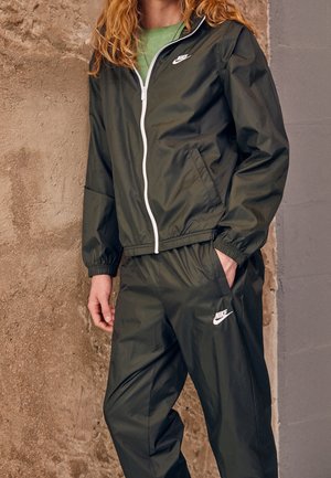 Personne portant un survêtement Nike vert foncé avec une fermeture éclair blanche et des logos, debout devant un mur texturé beige et gris.