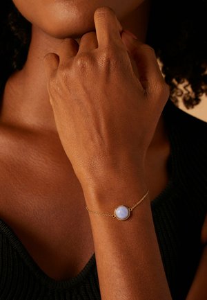 Gros plan sur la main d'une personne près du menton, portant un délicat bracelet chaîne en or avec une pierre ronde de couleur violet clair.