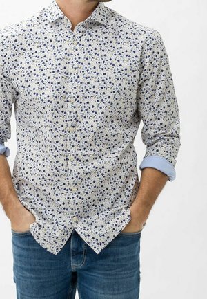 Man wearing a white buttoned floral shirt with blue and gray flowers, sleeves rolled up, and blue jeans with hands in pockets.