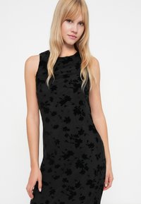 Zwarte mouwloze jurk met een bloemige fluwelen textuur en subtiele glans. Aansluitend ontwerp met geplooide details bij de halslijn en een soepele silhouet.