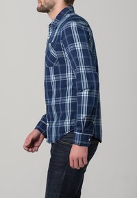 Košile s dlouhým rukávem v námořnické modré a světle modré kostkované vzoru, s límečkem na knoflíky, přední kapsou a kulatým lemem, nošena přes tmavé džínové kalhoty.