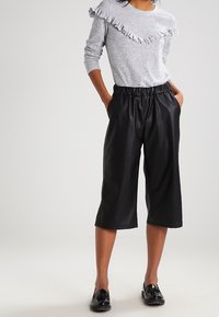 Maglia grigia a maniche lunghe con scollatura a balze, abbinata a pantaloni in pelle nera cropped. La modella indossa scarpe nere slip-on.