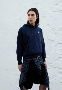 Μπλε ναυτικό βαμβακερό hoodie με μια μπροστινή τσέπη και λευκό λογότυπο, συνδυασμένο με μαύρο σορτς και μια δεμένη καρό πουκάμισο γύρω από τη μέση.