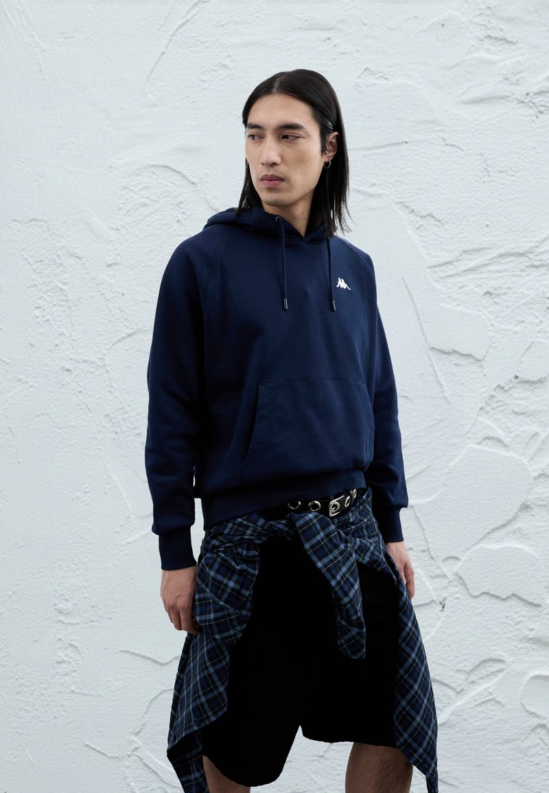 Μπλε ναυτικό βαμβακερό hoodie με μια μπροστινή τσέπη και λευκό λογότυπο, συνδυασμένο με μαύρο σορτς και μια δεμένη καρό πουκάμισο γύρω από τη μέση.