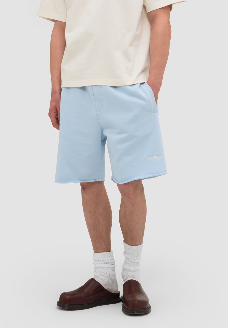 Personne portant un short bleu clair jusqu'au genou, des chaussettes blanches texturées, des chaussures marron sans lacets et une chemise crème à manches courtes, debout les mains dans les poches.