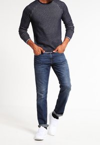 Pull en tricot bleu marine avec un motif à pois, associé à un jean en denim à lavage moyen. Chaussures blanches minimalistes, style baskets.