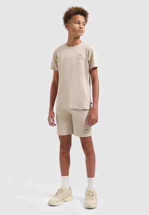 Teen wearing beige matching t-shirt and shorts, white socks, and beige sneakers, standing and looking to the side on a plain background.