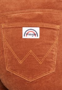 Pantalones de pana Wrangler en un cálido color naranja, con un bolsillo trasero que presenta una etiqueta blanca y un diseño de arco multicolor. Costuras amarillas acentúan el bolsillo.