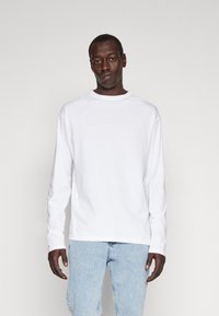 Mand iført en ensfarvet hvid langærmet skjorte og lyseblå jeans, stående mod en neutral grå baggrund.