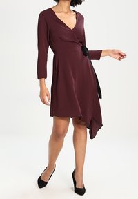 Bordowa sukienka kopertowa z dekoltem w kształcie litery V, rękawami o długości trzech czwartek oraz wiązaniem w talii. Posiada asymetryczny dół i miękką teksturę tkaniny.