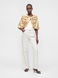 Femme portant une cape en satin doré, un haut blanc sans manches, un pantalon large blanc et des sandales noires à talons, debout avec une main dans la poche.