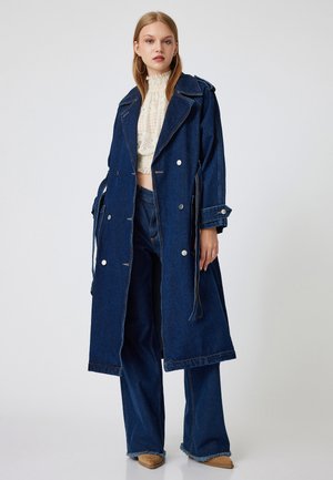 Lungo cappotto di jeans scuro con ampi revers, bottoni argentati e lacci regolabili. Indossato sopra un top di pizzo leggero e jeans svasati coordinati.