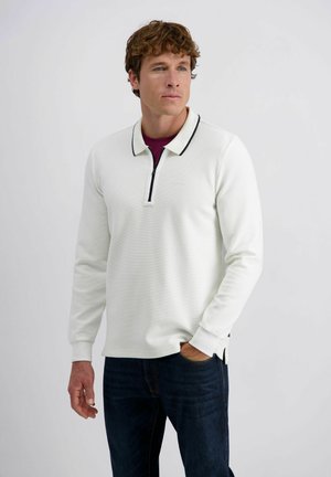 Man wearing white long-sleeve polo with black collar trim and dark jeans, standing with one hand in pocket against plain background.